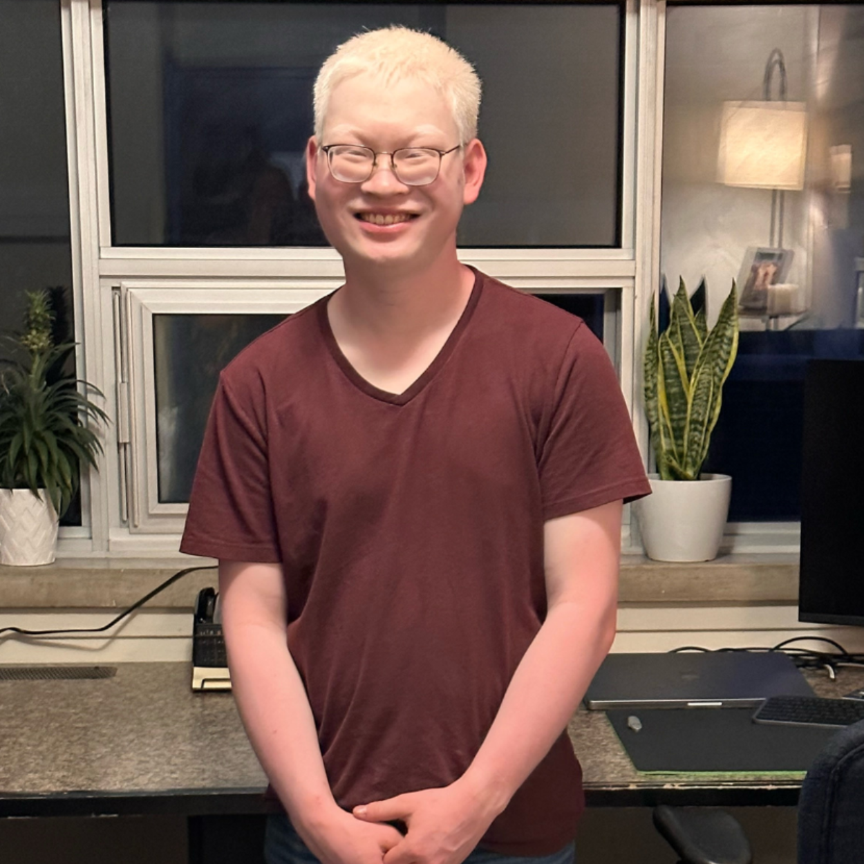 Adian, a young man with white hair, stands in-front of a home office/computer workstation. Aidan wears glasses and a brown t-shirt with jeans. He is smiling and standing with his hands clasped together.