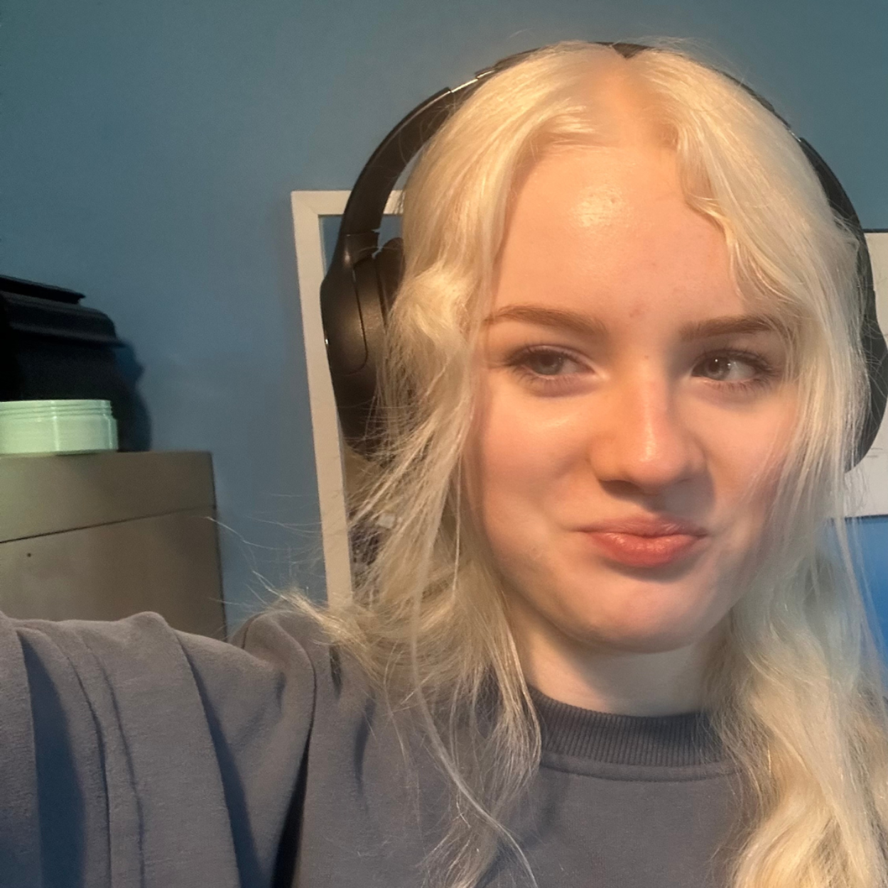 Acacia, a young woman with white hair, smiles and poses for a selfie-style photograph. She is wearing headphones over her ears and a long grey sweatshirt.