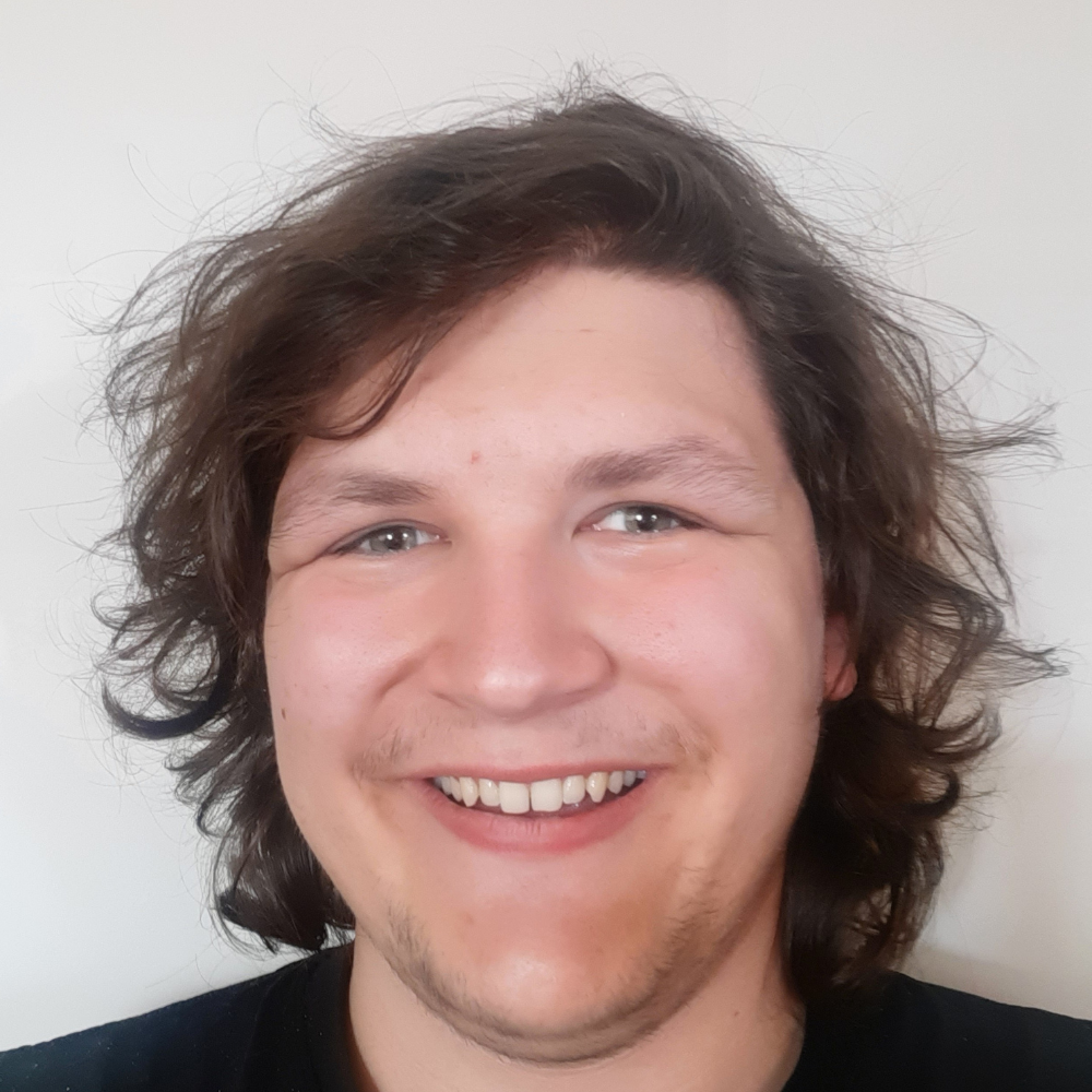 Tate, a young man with brown hair, poses for a selfie-style photograph. He is wearing a black t-shirt and is smiling.
