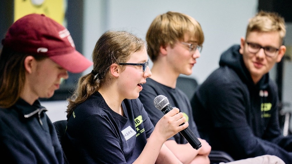Three youths, each with light skin, sit in a row. The young girl on the left speaks into a microphone.
